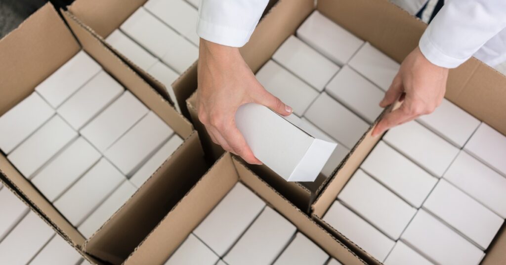 A person packs sealed white packages into a larger cardboard box. Other boxes with white packages are nearby.