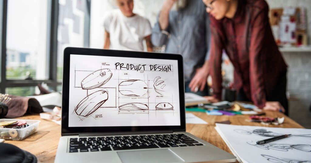 A group of designers stands together to brainstorm behind a computer with a product design mock-up on the screen.